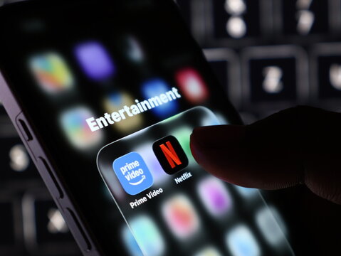 Closeup of a persons finger touching the netflix app icon on a smartphone screen, with the prime video app visible next to it in an entertainment folder