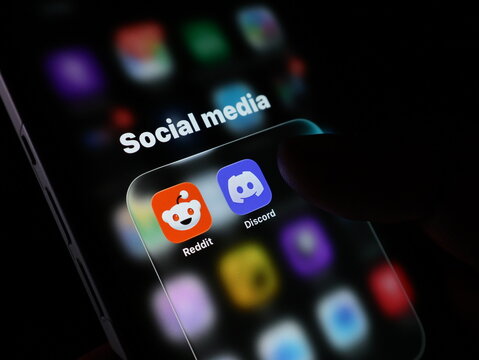 Closeup of a smartphone screen showing social media app icons including reddit and discord with a finger touching the screen in the dark