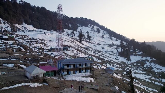 Aerial view of Chopta Tungnath with a telecommunications tower and small buildings on a snowy mountain slope in Chopta, Uttarakhand, India.