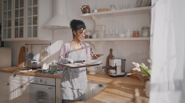 Joyful housewife or culinary blogger taking mobile photo of fresh baked cookies. Medium portrait of charming african american woman in apron holding baking sheet with homemade sweets in home kitchen