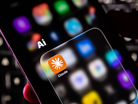 Closeup of a smartphone screen displaying the claude ai app icon within a folder labeled ai, held in a hand against a dark keyboard background
