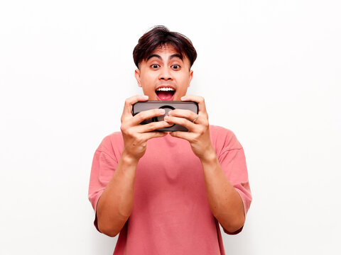 Excited young Asian man holding smartphone horizontally while playing mobile game with surprised expression, celebrating victory or high score on isolated white background.