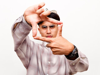 Naklejka premium Serious young Asian creative director making a finger frame gesture around his eyes, focusing closely at the camera to search for vision against a white studio background.