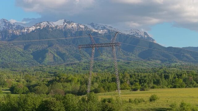 Impressive high tension electricity pylon with snow capped Bucegi mountains in background, gorgeous spring landscape, slow ascending aerial medium telephoto