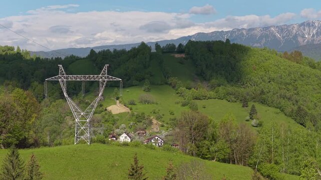 Imposing electricity pylon on a green hill with high voltage lines crossing a valley near Piatra Craiului mountains, dynamic cloud shadows, slow lateral aerial tracking