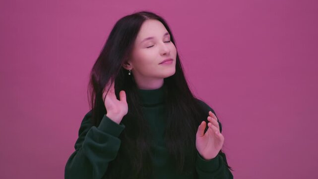 Teenage girl in a pink studio in a green sweatshirt shakes her long dark hair in a slow mo. Young brunette woman wiggles her locks of hair. Beautiful female, generation Z or alpha. Moving hairstyle