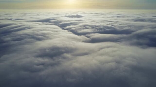Cinematic aerial view above dense white cloud layer with golden sunrise glowing on horizon during flight.