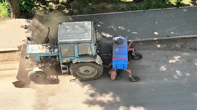 A blue tractor with a front attachment sweeps a dusty urban road. The machine is actively working, removing debris and maintaining cleanliness on a paved street.