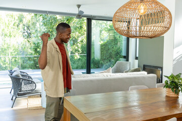 Fototapeta premium African American man standing at home by wood dining table raising fist, looking toward glass doors