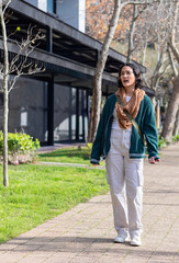 Fototapeta premium Adult woman walking along paved path beside building, holding phone and wearing green crossbody bag