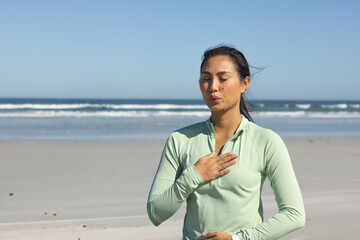 Obraz premium Mid-adult Asian woman practicing breathing at sandy beach in light green zip-up top, hands on chest
