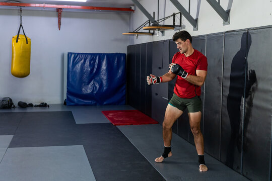 Man practicing striking in gym red-shirt MMA-gloves on mats near yellow heavy bag, copy space