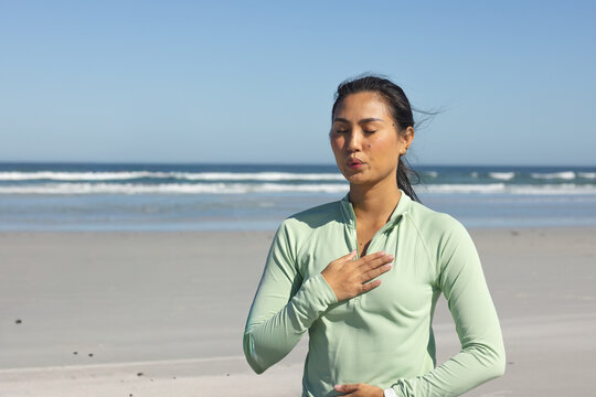 Mid-adult Asian woman practicing breathing at sandy beach in light green zip-up top, hands on chest