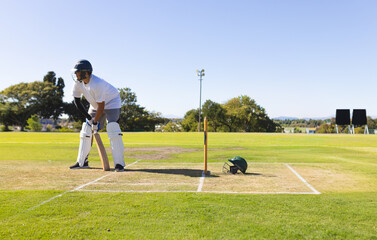 Obraz premium Male batter standing at crease by stumps on cricket ground holding wooden bat near spare helmet