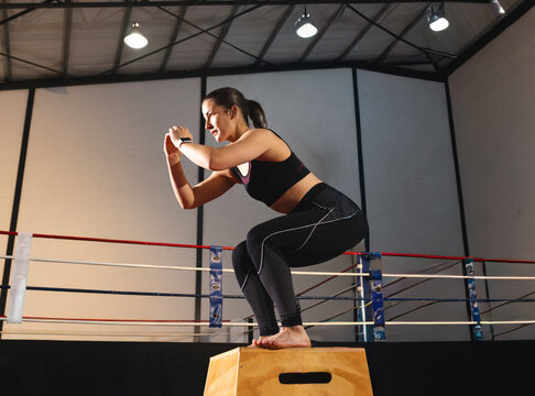 Female athlete holding squat on plyo box in boxing ring, wearing sports bra, wrist tracker