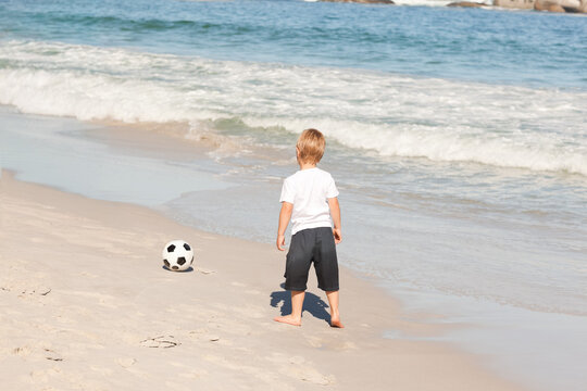 Boy child standing watching ocean at beach in white tee and dark shorts with soccer ball