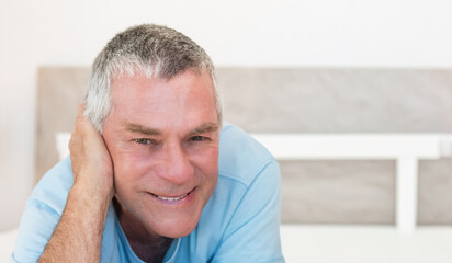 Senior man reclining on bed, propping left hand at face, wearing blue tee, headboard visible © wavebreak3