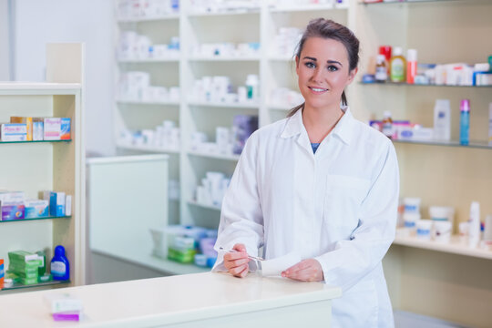 Woman pharmacist wearing lab coat working behind counter holding prescription and pen in pharmacy