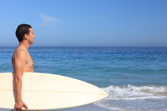Adult male standing at shoreline holding long white surfboard, barechested in trunks, copy space