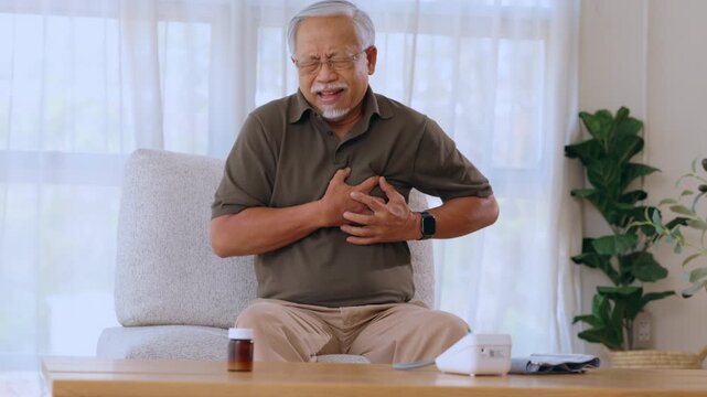 Senior Asian man sitting indoor on sofa at home experiencing severe chest pain and heart attack feeling distressed unwell in agony