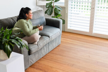 Obraz premium Mid-adult woman sitting crosslegged on gray sofa in green sweater holding gamepad in living room
