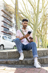Obraz premium Man sitting on bench scrolling smartphone and eating pastry with headphones at tree-lined street