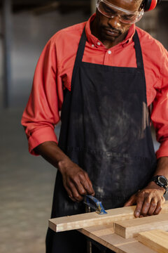 Middle-aged African American man in red shirt and apron smoothing timber at bench with hand plane