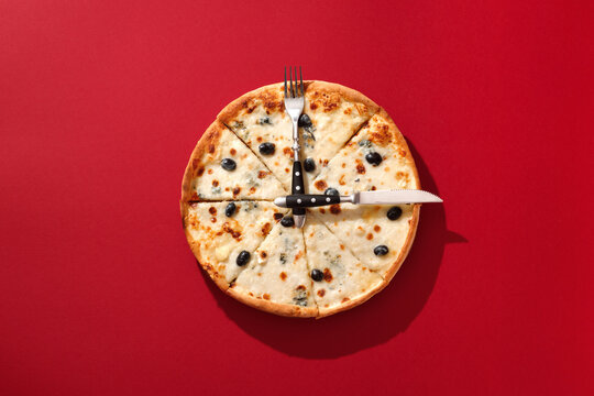 Pizza on a red surface in sunlight with fork and knife as clock hands. Creative and appetizing pizza clock concept for lunch or dinner time. Top view, flat lay.