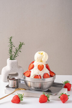 strawberry bingsu ice cream served in a silver bowl with slice strawberries on top and around it.served with a tray,spoon on the side and decorated with a white stone vase with a white base and a gray