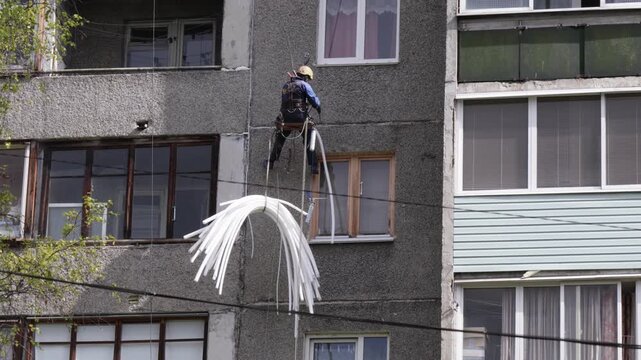 Worker suspended at height performs facade maintenance, applying insulation to seal gaps between concrete panels.