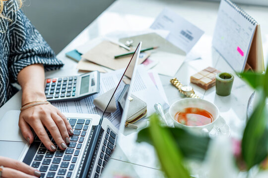Person typing on a laptop at a cluttered desk with tea and papers.