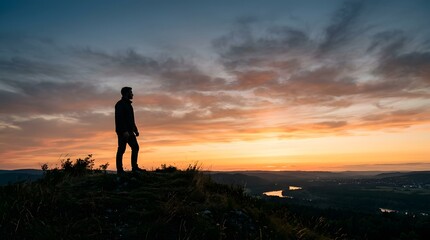 Fototapeta premium Lone hiker silhouetted against a dramatic sunset sky, overlooking a vast landscape with distant lights and a tranquil river.