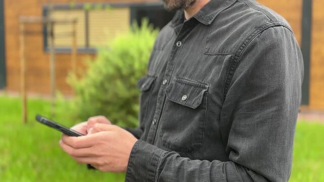 Adult man holding a smartphone outdoors near a green lawn, mobile communication, casual urban lifestyle, digital connection, everyday technology.