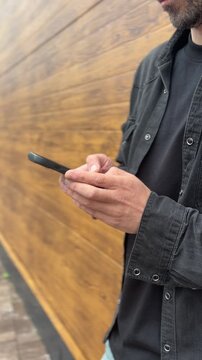 Adult man using a smartphone beside a wooden wall outdoors, mobile messaging, modern lifestyle, urban communication, casual technology use.