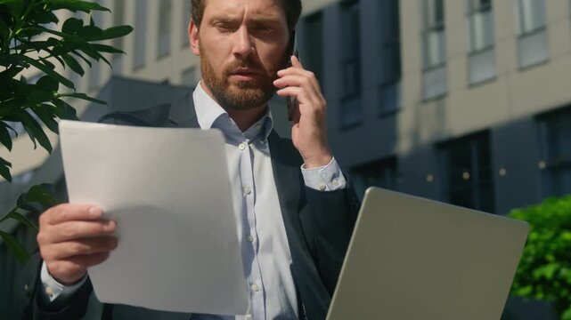 Serious focused Caucasian middle aged business man multitasking speaking smartphone holding documents in city busy male working laptop report discussing startup managing paperwork businessman outdoors