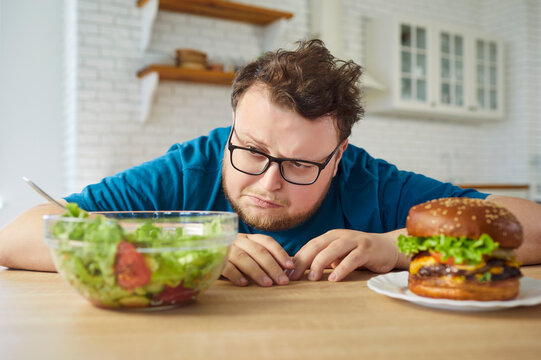 Overweight man faces burger and salad choice. At home, he weighs diet goals against temptation and questions his nutrition. Salad versus an unhealthy burger. Simple willpower and choice theme.