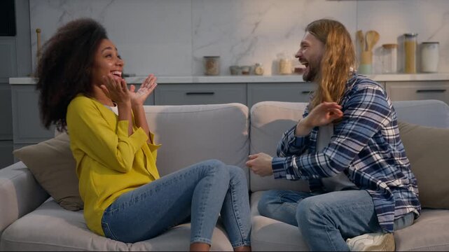 Multiethnic couple man woman talking at home couch sharing good news excited happy together Caucasian male African American female amazed gossip conversation love family friendship indoors diversity
