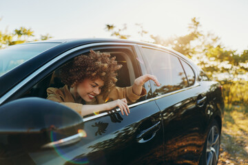 Fototapeta premium Smiling woman with curly hair leaning from a car window in a sunlit outdoor setting, capturing a candid lifestyle moment with retro film color and travel mood