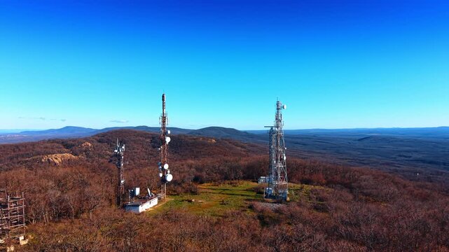 Close aerial view of telecom antenna towers on bare hilltop above rolling hills. Two telecom towers with dish antennas stand on a hilltop clearing amid bare brown scrubland and rolling hills beyond.