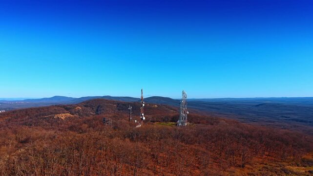 Aerial view of two telecom antenna towers on bare hilltop with rolling hills. Two tall antenna towers with dish arrays rise from a bare hilltop of brown leafless scrubland under a deep blue sky.