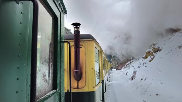 Old steam train travels through a snowy mountain landscape, belching smoke and steam in scenic winter motion.
