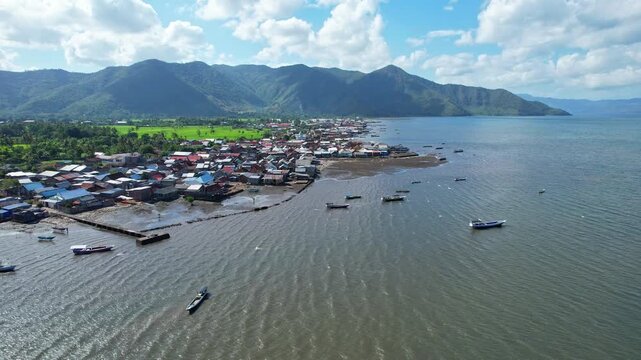 Aerial drone footage of Kempo shore city in Dompu regency, Sumbawa island, Indonesia, with wooden fishing boats at anchor, the bay with mountains behind, and green lush rice field, a mosque