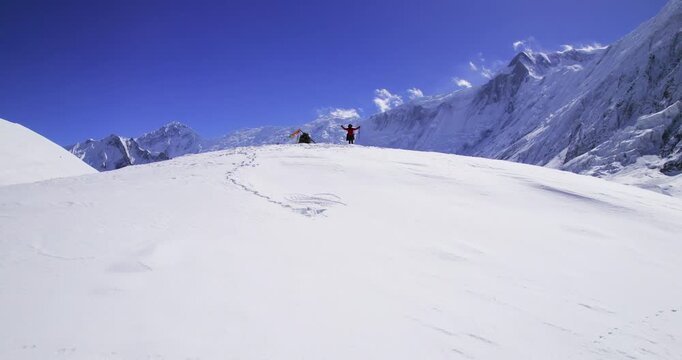 A triumphant trekker in a red jacket raises both arms with trekking poles beside prayer flags and a backpack at a snowy Himalayan camp in Nepal.