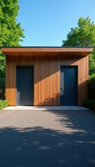 Fototapeta premium Modern wood building with two dark doors sits near green trees. An asphalt road with dappled shadow leads to its entrance. Clear blue sky is visible above.