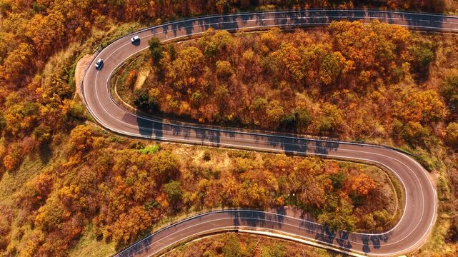 Aerial view of two tight hairpin road bends with cars in orange autumn forest. Two white cars navigate separate U-shaped hairpin bends on an orange autumn hillside as tree shadows stripe the asphalt.