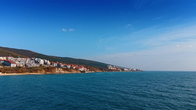 Drone lateral view of coastal resort town on sandy cliffs above the Black Sea. White apartment buildings top sandy coastal cliffs as an autumn forested hill rises behind the seaside resort town.