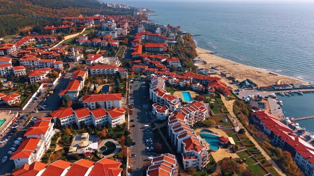 Close aerial view of beachfront resort complex with pools streets and marina. Red roofed resort apartments surround pools and gardens as a sandy beach and marina jetty meet the sea on the right edge.