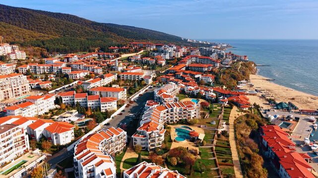 Aerial view of dense beachfront resort complex with red tile roofs and pools. White resort apartments with terracotta roofs surround pools and gardens as a sandy beach lines the sea on the right.