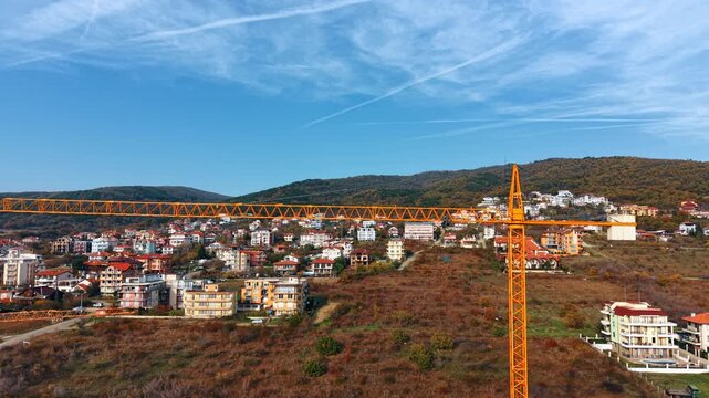 Aerial view of coastal town with tall orange construction crane in foreground. An orange tower crane rises above a hillside coastal town as late autumn scrub covers the slope below forested hills.