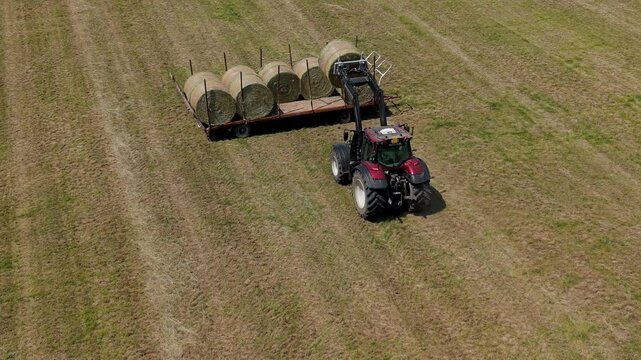 Tractor Harvesting Hay Bales: A red tractor in a dry grassy field uses a front loader to lift hay bales onto a trailer. The aerial view captures the agricultural process under a bright, clear sky.